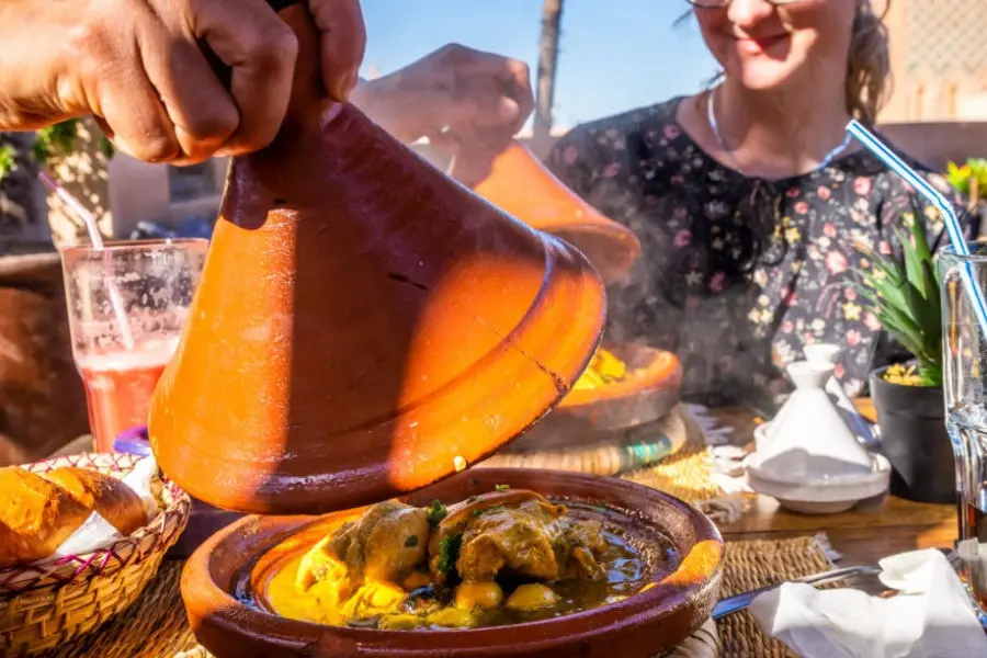 A person lifts the lid of a traditional Moroccan tagine pot, revealing steaming food, while a woman smiles in the background at an outdoor restaurant.