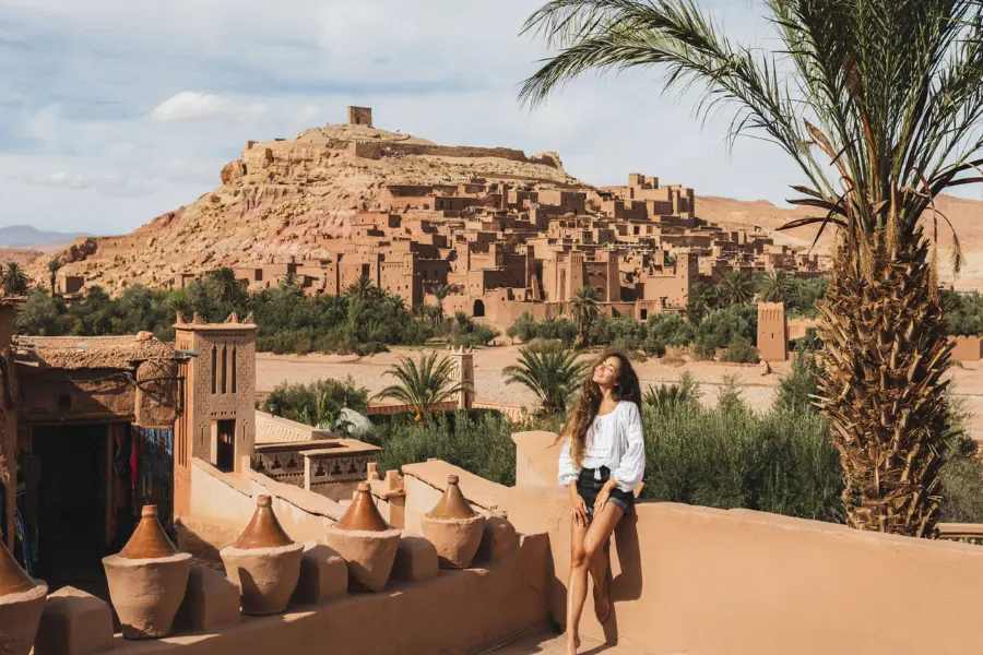 A woman sits on a terrace in the sun, smiling, with a palm tree beside her and an ancient desert fortress village in the background.