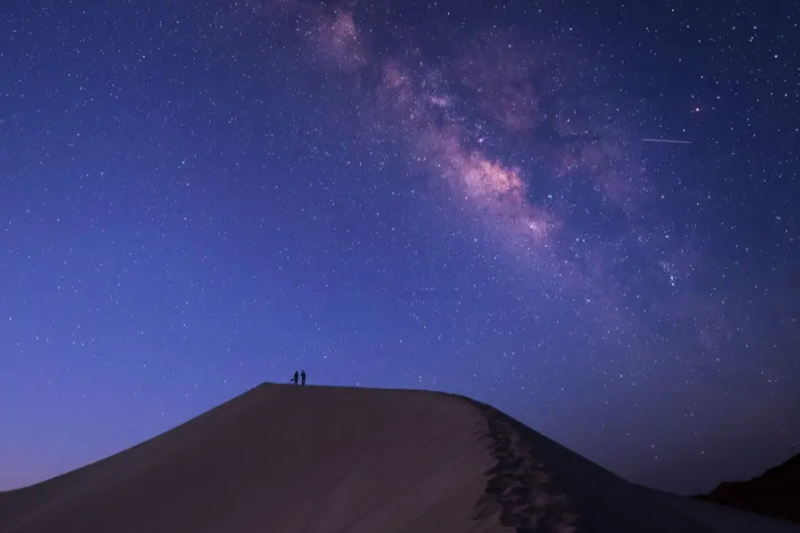 Two people stand on top of a sand dune at night beneath a vivid Milky Way sky filled with stars.