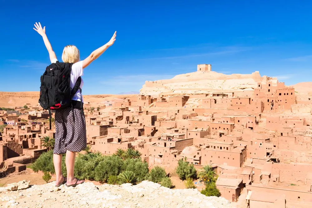 A female traveler with a backpack stands on a cliff with arms raised, overlooking an ancient desert village made of mud-brick buildings under a bright blue sky.